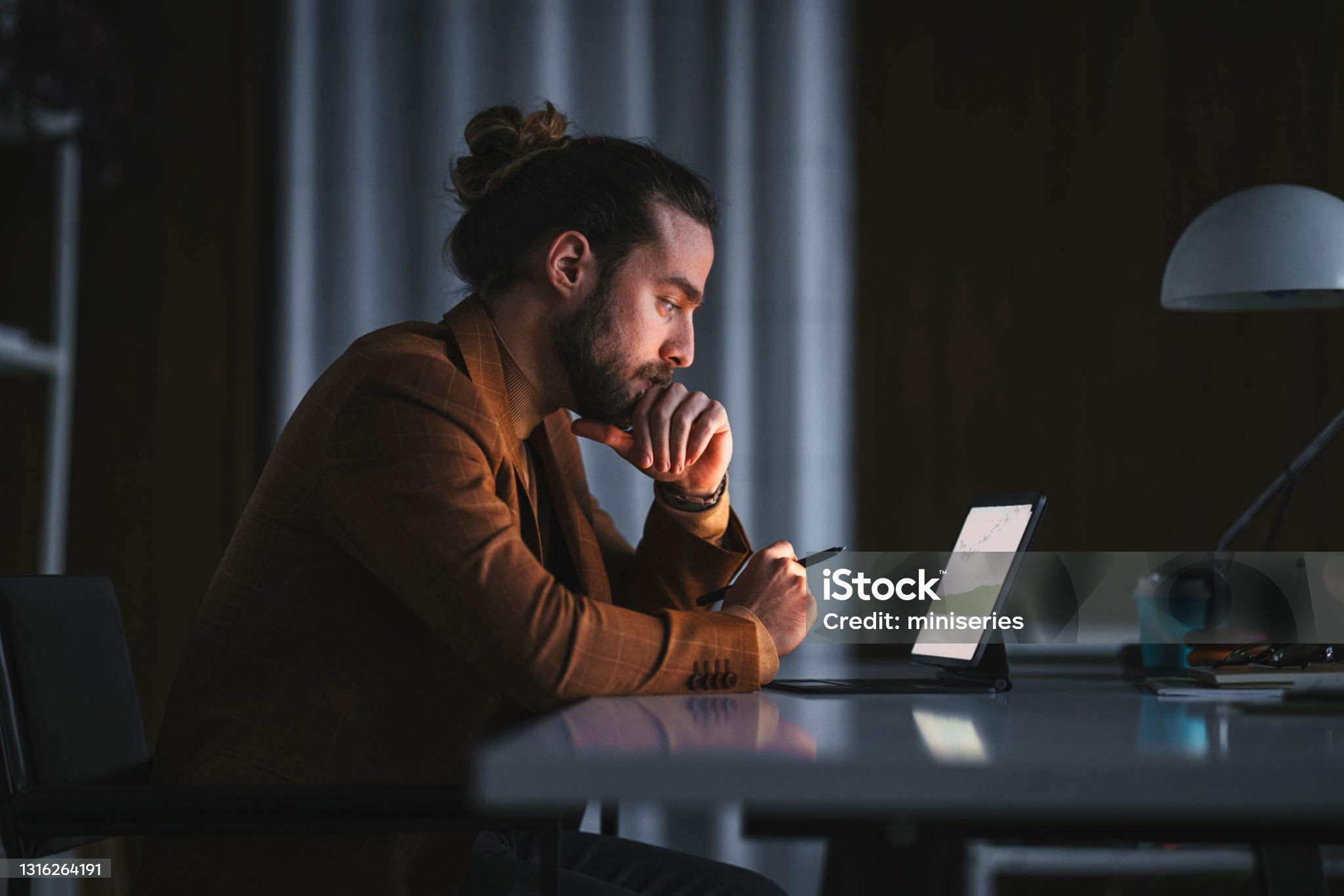Side view of serious thoughtful adult bearded male in stylish wear sitting at table with modern laptop and analyzing business information while working in office in late evening